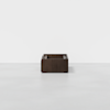 A minimalist dark wooden box with open sides and cut-out handles sits centered on a plain white floor against a white background.