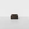 A minimalist dark wooden box with open sides and cut-out handles sits centered on a plain white floor against a white background.