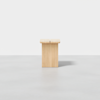 A minimalist wooden stool or side table with a light natural finish, positioned on a smooth, light gray floor against a plain white background.