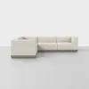 A modern, L-shaped sectional sofa with light beige upholstery and a wooden base, placed in an empty, minimalist room with a light gray floor and white walls.