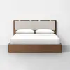 A modern wooden platform bed with a light upholstered headboard, white bedding, and two white pillows, set against a plain white wall and gray floor.