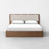 A modern wooden platform bed with a light upholstered headboard, white bedding, and two white pillows, set against a plain white wall and gray floor.