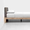 A minimalist wooden bed frame with a gray upholstered headboard and white bedding, including a pillow and sheets, positioned against a plain white wall and floor.