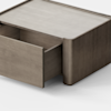 A minimalist wooden storage box sits on a light floor, with one drawer partially open, revealing its simple, clean-lined design against a plain white background.