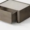 A minimalist wooden storage box sits on a light floor, with one drawer partially open, revealing its simple, clean-lined design against a plain white background.