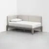 A modern daybed with a light wood frame, beige upholstered backrest, white mattress, and a single white pillow, placed in the corner of a minimalist room with gray flooring and white walls.