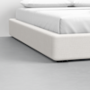 A close-up view of a modern bed with a light beige upholstered frame and white bedding, set on a smooth, light gray floor in a minimalistic room.