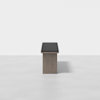A minimalist, modern bench with a dark cushion and a rectangular wooden base, centered in a white, empty room with a smooth floor.