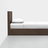 A simple, modern wooden bed with a tall headboard, neatly made with white bedding and a single white pillow, positioned against a white wall on a grey floor.