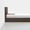 A simple, modern wooden bed with a tall headboard, neatly made with white bedding and a single white pillow, positioned against a white wall on a grey floor.