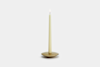 A single lit cream-colored taper candle stands upright in a simple, round brass holder against a plain light gray background.