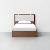 A single bed with a wooden frame and light-colored upholstered headboard, made up with white sheets, blanket, and pillow, set against a plain white wall and gray floor.