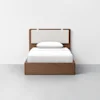 A single bed with a wooden frame and light-colored upholstered headboard, made up with white sheets, blanket, and pillow, set against a plain white wall and gray floor.