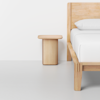 Minimalist bedroom scene with a light wood bed frame, crisp white bedding, and a matching light wood bedside table against a plain white wall and light gray floor.