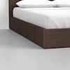 A dark brown wooden bed frame with white bedding and pillows sits on a light gray floor in a minimal, modern room.