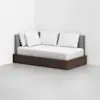 A modern daybed with a dark wooden base, gray upholstered backrest, and four white pillows, set against a plain white corner in a minimalistic room.