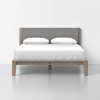 A modern bed with a light wooden frame, a gray upholstered headboard, white bedding, and two white pillows, set against a plain white wall and floor.