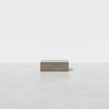 A minimalist, rectangular wooden box sits centered on a smooth, light gray floor against a plain white background.