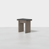 A modern wooden stool with a rectangular black cushioned seat, featuring simple, chunky legs and a minimalist design, placed on a light gray floor against a plain white background.