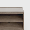 A close-up of a wooden TV stand or shelf unit with a light brown finish, featuring an open shelf and a small circular cable management hole at the back. The background is plain white.