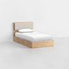 A single wooden bed with a padded beige headboard, white bedding, and a pillow, set against a plain white wall in a minimalistic room with a light gray floor.