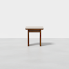 A small wooden stool with a light-colored cushioned seat stands on a plain, light gray floor against a white background.