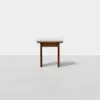 A small wooden stool with a light-colored cushioned seat stands on a plain, light gray floor against a white background.