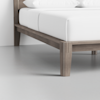 Close-up view of the corner of a wooden bed frame with light brown legs, a white mattress, and white bedding on a smooth, light gray floor against a white wall.