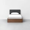 A modern bed with a dark gray upholstered headboard, white bedding, and a wooden frame sits on a light gray floor against a plain white wall.
