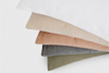Four textured pillows in beige, brown, gray, and olive green are stacked in a fan shape beneath a white pillow, all on a white background.