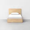 A wooden bed frame with a headboard and footboard, topped with a neatly made white bed and a single white pillow, set against a plain light-colored wall and floor.