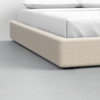 Close-up of a beige upholstered bed frame with a white mattress and pillows, positioned on a light gray floor in a minimalist bedroom setting.