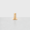 A small, minimalist wooden sculpture stands centered on a smooth, light gray surface against a plain white background. The sculpture is rectangular with simple, clean lines.