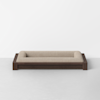 A modern pet bed with a low, dark wooden frame and a rectangular beige cushioned surface, placed against a plain white wall on a light gray floor.