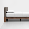 A minimalist bed with a wooden frame, a gray upholstered headboard, and white bedding and pillow, set against a plain white wall and floor.