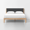A minimalist bed with a light wood frame, dark upholstered headboard, two white pillows, and a white duvet, set against a plain white wall and floor.