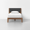 A single wooden bed with a dark gray upholstered headboard, white bedding, and a white pillow, set against a plain white background.