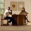 Two people sit at a wooden dining table with mugs and plates, facing the camera. Behind them are two abstract art pieces on the wall and a large window with trees visible outside.