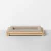 A modern rectangular pet bed with a light wood frame and beige upholstered cushion, placed on a smooth, light gray floor against a white wall.