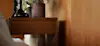 A close-up of a wooden side table with a drawer, featuring a potted plant and a dark glass vase on top, set against a wooden wall in a warmly lit room.