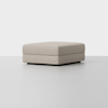 A large, square, light beige ottoman with a simple, modern design sits on a smooth, light gray floor against a plain white background.