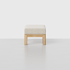 A minimalist ottoman with a light-colored cushioned seat and a natural wood frame, set against a plain light gray background.