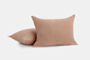 Two light brown textured pillows are shown against a plain white background; one pillow is standing upright while the other is lying down behind it.