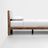 A minimalist wooden bed frame with a light wood finish, white upholstered headboard, white pillow, and neatly made white bedding, positioned against a plain white wall and floor.