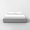 A modern bed with a light gray upholstered base, white mattress, and two white pillows, set against a plain white wall in a minimalist room.