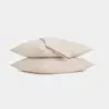 Two beige pillows stacked on top of each other with a matching folded sheet placed on top, set against a plain white background.