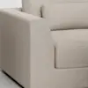 Close-up view of a modern beige fabric sofa featuring a thick seat cushion, rectangular backrest, and wide armrest on a light-colored floor.