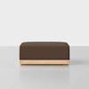 A square, brown upholstered ottoman with a wooden base, placed on a smooth, light gray surface against a plain white background.