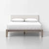 A neatly made bed with a light upholstered headboard, two white pillows, white bedding, and a wooden frame, set against a plain white wall and gray floor.