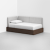 A modern daybed with a dark wood base and light gray upholstered back and side panels, fitted with a white mattress and pillow, positioned in the corner of a minimal, white-walled room.